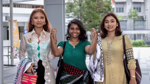 Provided by Kokila Annamalai Three women dressed in clothing in the colours of the Palestinian flag - red green and black - while wearing keffiyeh scarves hold hands and smile at the camera