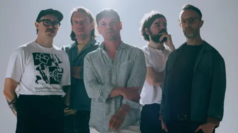 Prescription PR Five men looking into the camera, with light beaming down behind them. Two of the group are wearing glasses and all of them wear serious expressions on their faces. 