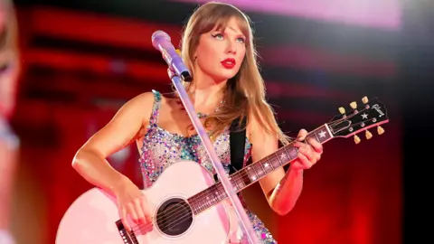 Getty Images Taylor Swift is on stage in a glittering, multi-coloured dress and holding a white guitar decorated with stars. She is wearing bright red lipstick and looking out across a crowd from behind a silver microphone and stand, in front of a blurred stage which is visibly illuminated in red.