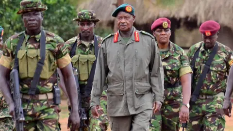 BBC President Yoweri Museveni, in a green beret and khaki uniform, surrounded by members of the  Special Forces Command (SFC) in green camouflage fatigues. Two are wearing maroon berets and two in floppy hats are armed with automatic rifles.