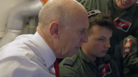 BBC Defence Secretary John Healey in side profile, wearing a white shirt and red tie, alongside RAF crew in uniform 