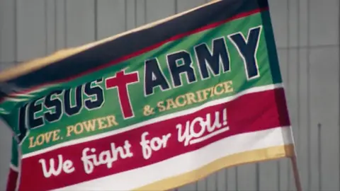 DocsvilleStudios/BBC Archive A green and red Jesus Army flag which reads: 'Love, Power & Sacrifice. We fight for you!'. The flag is blowing in the wind. 