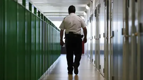 Getty Images A prison guard walks through a cell area at HMP Berwyn. He is walking away from the camera, passing white cells doors on his right hand side and a tall green railing to his left.