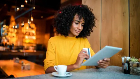 Getty Images A woman wearing a yellow jumper holds her credit card in one hand while she looks at a tablet in a cafe. 