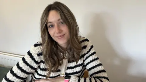 Abbi Lily Abbi, a young woman with dark blonde hair. She is wearing a black and white striped cardigan and is sitting at her kitchen table