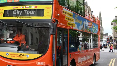 Getty Images A red open-top city tour bus in Edinburgh. The buildings of the Royal Mile can be seen in the background. There are a few passengers on the top deck of the bus, and a man in an orange hi-vis in the driver's seat. 