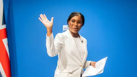 PA Media Kemi Badenoch wearing a white jacket and white t-shirt is pictured waving to the camera. There is a union jack pictured to the left.