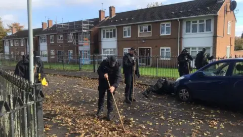 BBC Six police officers in black uniform and caps searching a residential street. Two officers are in the road holding wooden poles to search through the leaves on the floor. A dark blue car is parked on the right. One officer is looking under the car and two others are stood next to it. The other officer is on the left looking at the pavement. Two-storey houses are in the background, one has scaffolding up. 