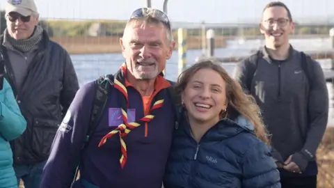 A man with embracing a woman from the side. They are shoulder-to-shoulder and looking towards the camera. The man has a goatee and has a neckerchief around his neck, and is wearing white-framed sunglasses on his head.