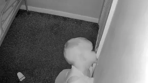 Jess Harry A black and white still image from a baby montiro of a young boy biting on a white doorframe. Behind him is his metal bedframe.