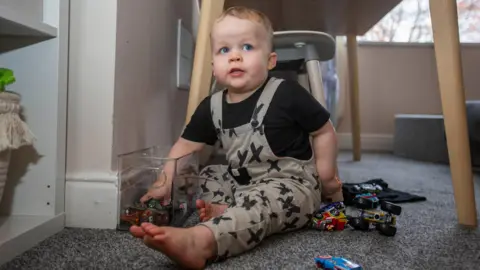 Craig Colville A little boy sat on grey carpet under a table. He is surrounded by toy cars and has his hand in a plastic tub of toy cars, with a cheeky expression on his face.