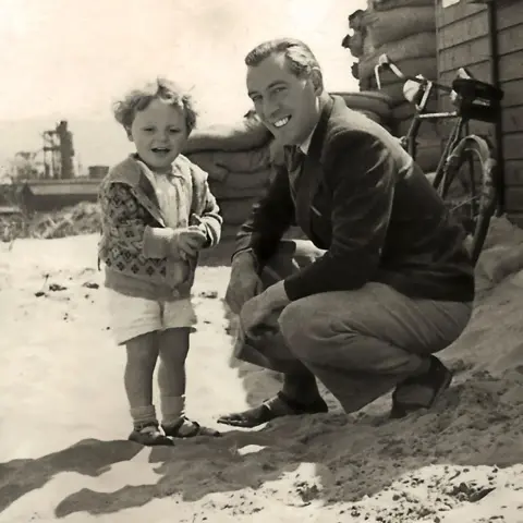 Sir Anthony Hopkins Anthony at 3, smiling, in white shorts and embroidered cardigan, curly hair, and 
his father, Richard, crouching in beige trousers and dark jacket
at Aberavon Beach in 1941 