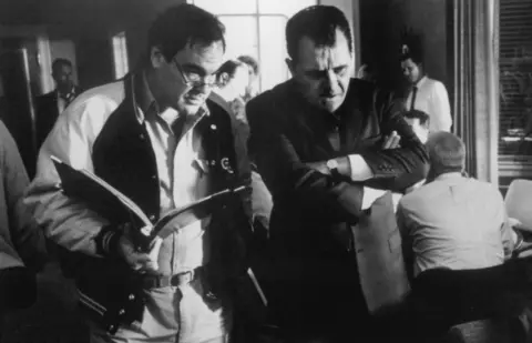Hollywood Pictures Behind the scenes black and white photo showing:
The director, Oliver Stone (L) with glasses and holding a file  talking to Hopkins in character in a suit with his arms closed
