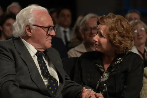 Warner Bros. Side profiles of Hopkins as Sir Nicholas Winton in dark grey suit and tie with gold circles. looking at & holding the hand of woman in black jacket and black, grey and white scarf
