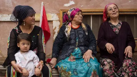 Getty Images A young Uyghur women,  with a small child on her knee, sits next to two older women on a street in Kashgar City, Xinjiang. All three are dressed in traditional garments with head scarves