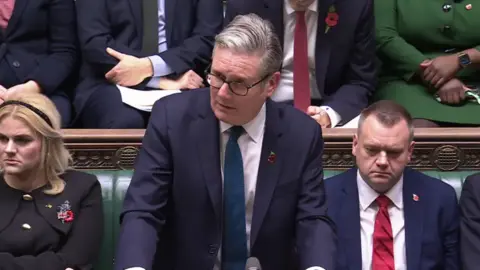 Parliament TV Keir Starmer at the despatch box in the House of Commons. He is wearing a blue suit and has a poppy on his lapel. He is flanked by two other MP who are sitting on the green benches behind him. 