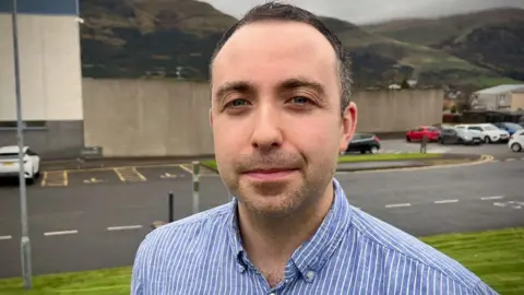 Head and shoulders shot of Glenochil GP Dr Jack Kildare. He has short black hair, stubble and is looking directly at the camera with a serious expression on his face. A prison building and parked cars can be seen in the background.