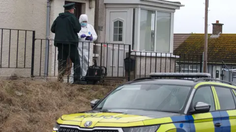 Pacemaker A police man and person in white PPE gear are standing outside a white pebble dashed house. A police car is parked in the foreground