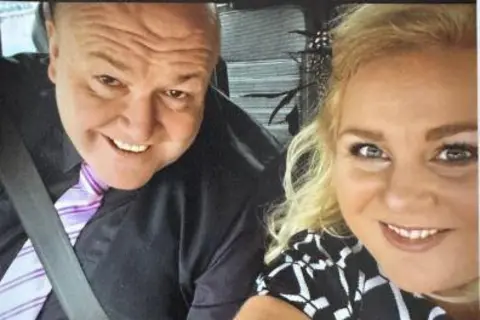 Family photo A man and woman in a car take a selfie together. The man is on the left and smiles. He is in a car as he has a seatbelt on. He has a black shirt, waistcoat and purple tie. A woman is next to him, she has blonde hair and a black and white dress on. She also smiles. It is a head and shoulders shot of the two of them. 