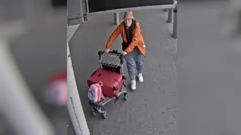 Sussex Police A man with a bucket hat and orange jacket leaves an airport wheeling a trolley with luggage on it. 