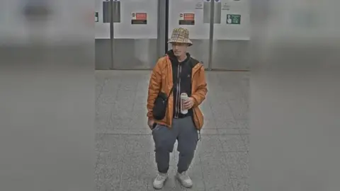 Sussex Police A man in an orange jacket with a bucket hat. He is drinking a pint can of Stella. 