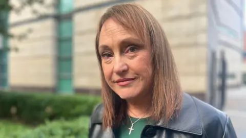 A woman, pictured from the shoulders up, looking at the camera. She is wearing a green told and black leather jacket. She has shoulder length brown hair. 