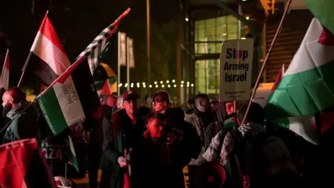PA Media Pro-Palestinian campaigners stage a demo outside Villa Park. Many are carrying flags and waving them. There is a banner that says "stop arming Israel". The stadium building is behind them with the lights on inside.
