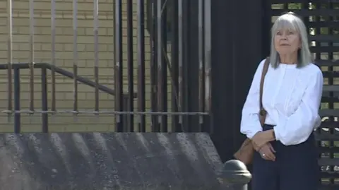 Two women leaving a black turntable style gate. The woman on the left is Anne Broughton who is wearing a white blouse and navy trousers. She has a brown bag on her left shoulder and has straight grey hair to her shoulders and a fringe. The other woman is standing side on and has grey short hair and is all in black. Her face is obscured. She has a black backpack on. 