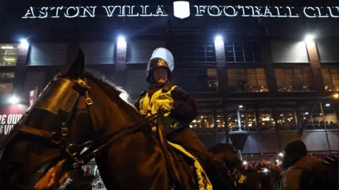 Reuters A police officer sits astride a horse which is in the foreground of the image, the ground is behind and the lit-up sign reading Aston Villa Football Club is in the background