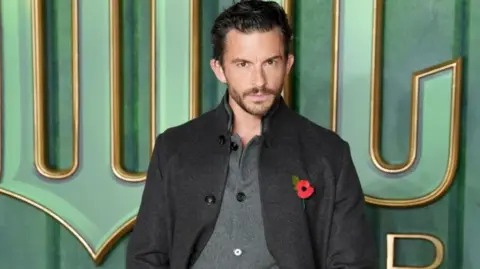 PA Media Actor Jonathan Bailey, who reprises his role of Prince Fiyero in the Wicked sequel, is pictured at the film's London premiere looking at the camera. He is wearing a dark grey jacket and Remembrance Day poppy over a grey buttoned top.