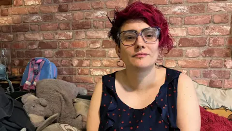 A woman with short pink hair, wearing glasses, heart-shaped earrings and a navy and red patterned top, sits on a sofa. Behind her is a brick wall. On the sofa are cushions, an elephant cuddly toy and a small blue and pink backpack.