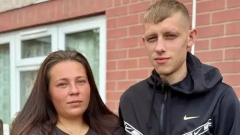 Anna Crossley/BBC Headshots of two people. On the left a woman with long dark brown hair wearing a black coat and a gold necklace. She is standing next to a taller man  who has short light brown hair and is wearing a dark blue sports top with a hood