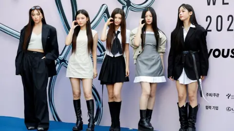 Reuters Members of NewJeans stand in a row at an event, posing for photos in front of a giant sign. They are dressed in black and white clothes.