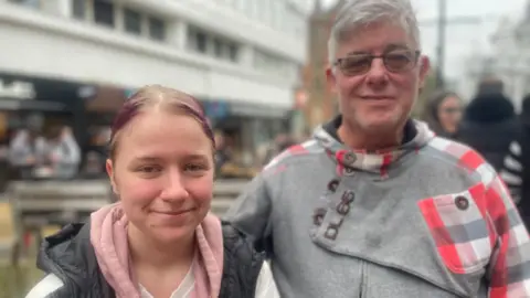 Simon Thake/BBC A man with grey hair and glasses and wearing a grey sweatshirt stands next to a younger woman with purple hair tied back on her head.
