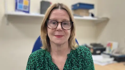 Maria Kane sat in an office. She is wearing a green leopard print shirt and has on glasses. Behind her is a desk with paper work on and a shelf.