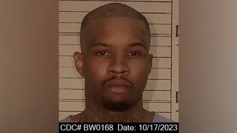 California Department of Corrections and Rehabilitation A mugshot showing rapper Tory Lanez. He has neck tattoos, a mustache and goatee beard. She is standing in front of a wall with black height markings. 