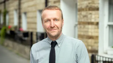 Elmwood Ivan Mato wearing a tie and button-down collar looks into the camera.