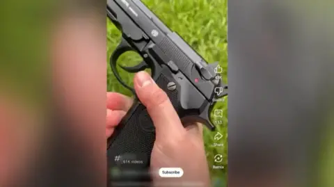 TikTok A screengrab from a TikTok video showing a hand holding a black handgun with grass in the background.