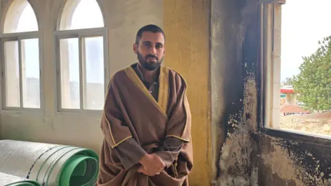 Imam Ahmad Salman. He is wearing a brown robe with yellow detail. He is standing next to a burnt wall where the paint has been blackened. The window next to it is empty of glass.
