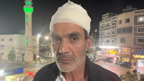 Hamad al-Jagoub abu Rabia. He has a bandage wrapped around his head and a grey stubble beard. Some dark hair can be seen poking out from beneath the bandage and he has thick, dark eyebrows. Behind him are some buildings, including shops and a tower lit up in green.