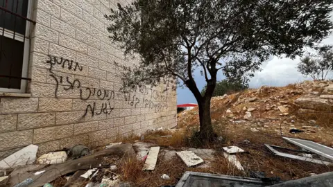 Hebrew graffiti on a white brick wall. In front of it is a tree surrounded by bit sof rubble, window frames and other detritus