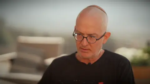 Amichai Luria. He has a shaved head with an orange yarmulke and is wearing black rimmed glasses. He is wearing a black t-shirt 