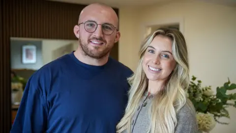 A man and woman stand close together both smiling into camera. The man has a shaved head, is wearing glasses and a dark blue t-shirt. The woman has long blonde hair, blue eyes and is wearing a grey cardigan.