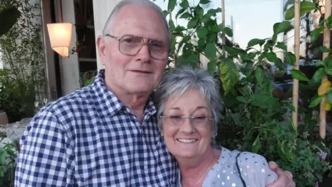 Submitted A man with a blue check-patterned shirt, receding white hair and glasses is standing with his arm around a woman, who has short grey hair and glasses and wearing a white shirt with silver sequins.