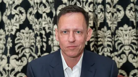 Nordin Catic/Getty Images for The Cambridge Union Peter Thiel wearing a white shirt and blue jacket in front of a floral wall