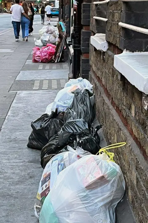 Loïc Frémond Rubbish bags lined along a pavement next to a brick building, including black and clear plastic bags filled with mixed waste. More bags, including pink ones, are visible further down the street near stacked cardboard. Several people are walking in the background.