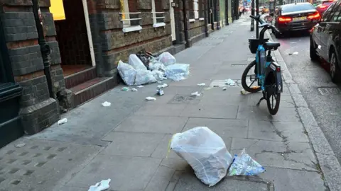 Loïc Frémond Rubbish from split bin bags strewn across a pavement next to a brick building, with several clear bin bags lined up along it. A blue rental e-bike is parked by the curb, and cars are on the road with traffic lights visible in the distance.
