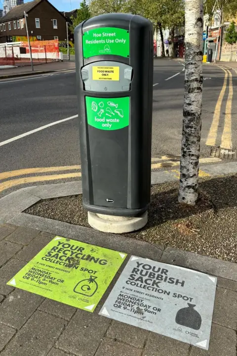 ReLondon A tall black bin for food waste positioned on a pavement beside a tree and a road. Two vinyl stickers mark collection spots for bin bags on the pavement in front of the bin, one for recycling sacks and one for rubbish sacks, with collection day information.