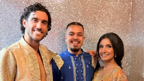 Josh Kapur Three people standing in front of a gold glitter wall. The man in the middle is wearing a blue outfit and the man and woman on either side of him are dressed in gold outfits. They are all smiling. 