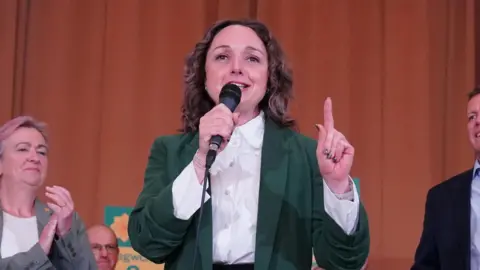 PA Media Woman in green jacket speaking into a microphone, behind her is a crowd holding Plaid Cymru signs. 